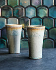 Two ceramic Pewabic Pints with a textured, glossy, ice-blue Frost glaze surface on a dark surface with a patterned Aurora Iridescent tile background.