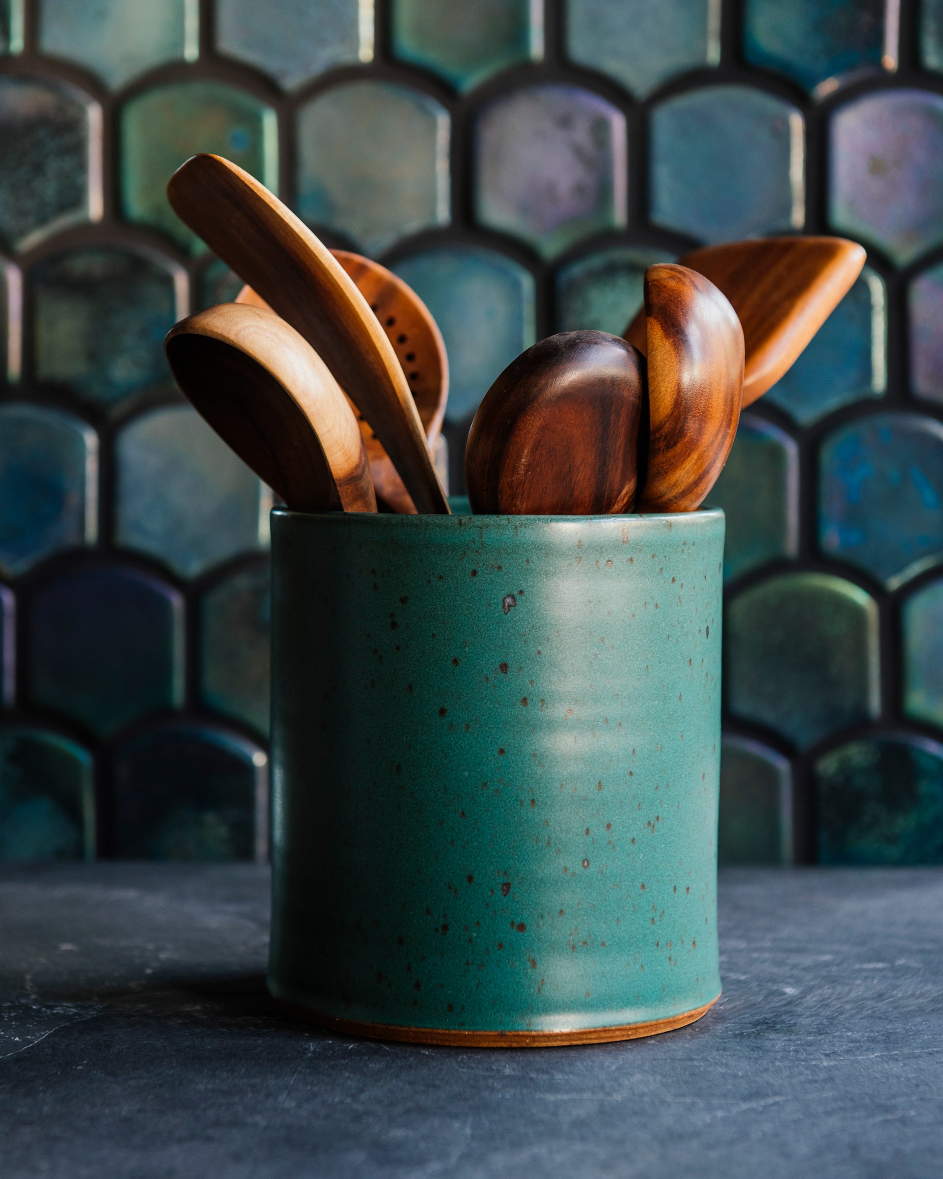 Floral arrangement in a freckled Greenstone vase with teakwood utensils against an Aurora Iridescent tile wall. 