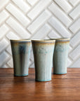 Three ceramic Pewabic Pints with a textured, glossy, ice-blue Frost glaze surface on a wood surface with a patterned Alabaster tile background.