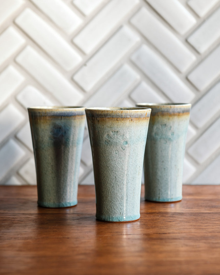 Three ceramic Pewabic Pints with a textured, glossy, ice-blue Frost glaze surface on a wood surface with a patterned Alabaster tile background.
