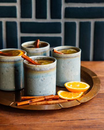 Four ceramic Rocks Cups with a warm beverage, cinnamon sticks, and orange slices on a wooden tray against a dark tiled wall.