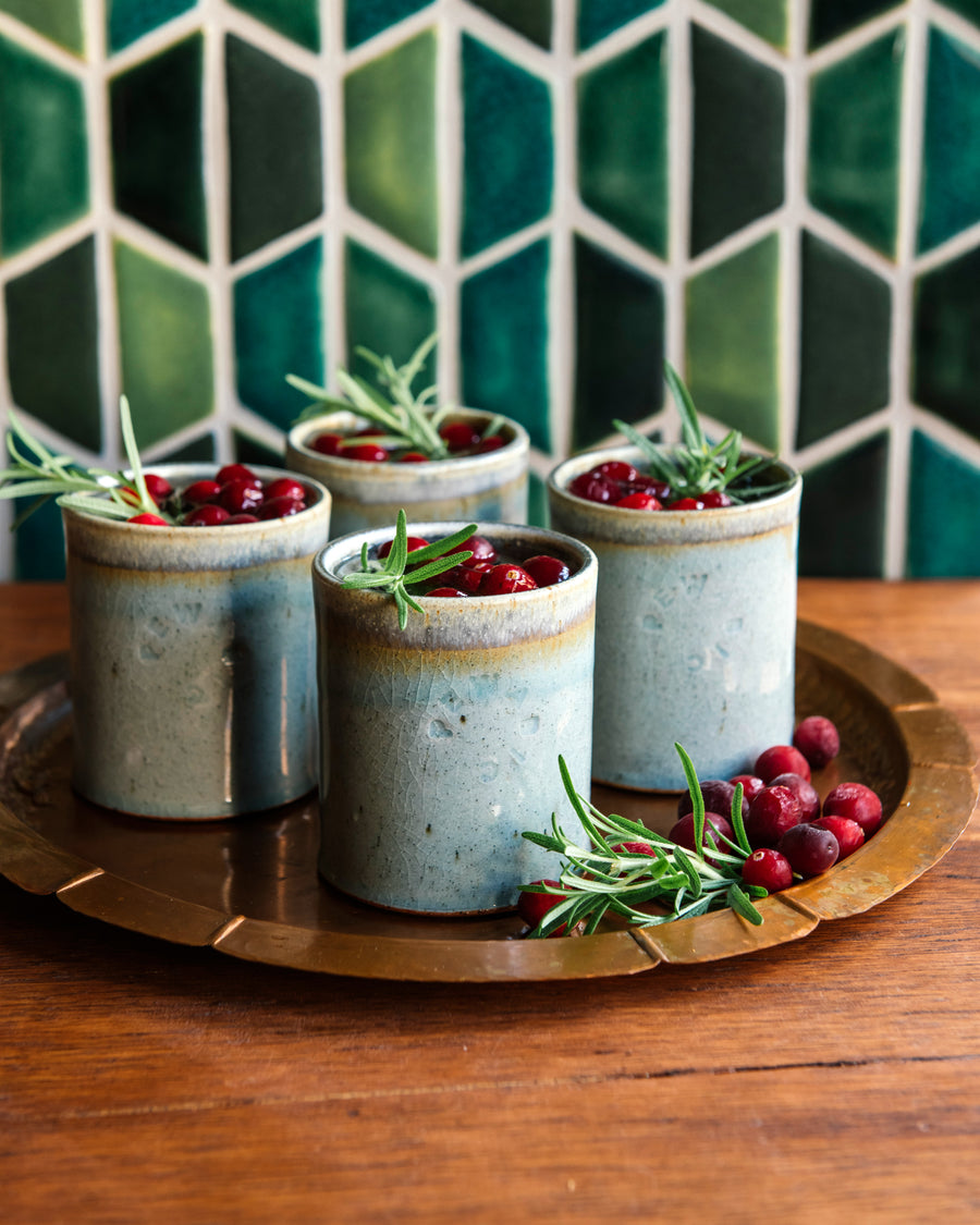 Five ceramic Rocks Cups with greenery and berries on a wooden tray against a geometric-patterned wall.