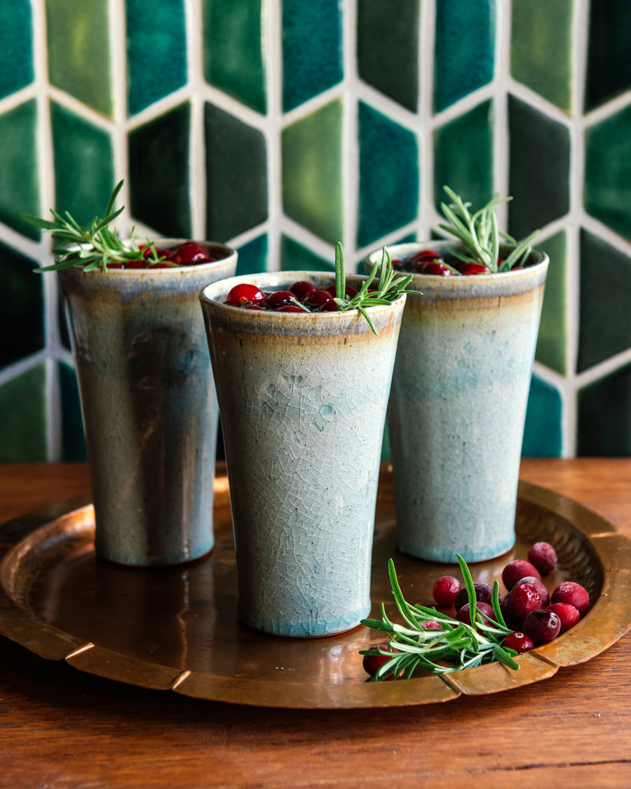 Three ceramic Pewabic Pints with a textured, ice-blue Frost glaze, garnished with rosemary and cranberries, on a wooden tray against a green half-hexagonal tiled wall.