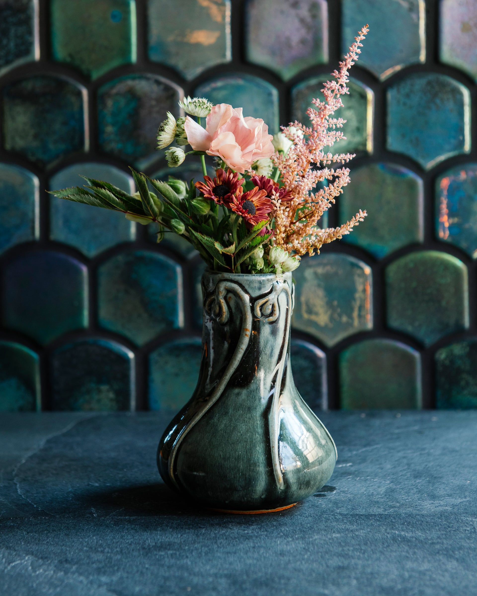 Glossy, deep green Balsam Snowdrop vase with blush-pink and vibrant green flowers against an Aurora Iridescent tile wall. 
