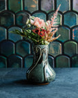 Glossy, deep green Balsam Snowdrop vase with blush-pink and vibrant green flowers against an Aurora Iridescent tile wall. 