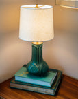 A matte green Elm glazed table lamp with an off-white shade on a stack of books against a neutral wall.
