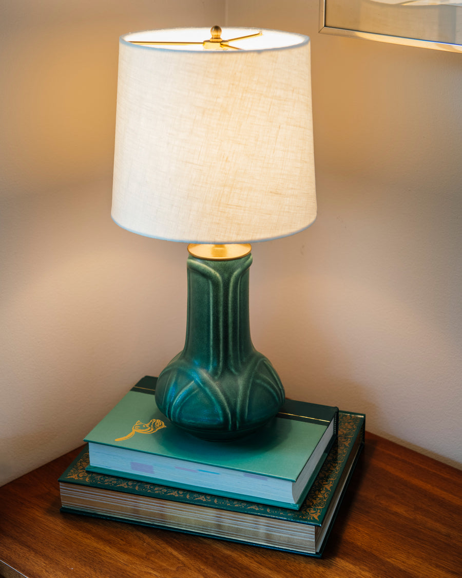 A matte green Elm glazed table lamp with an off-white shade on a stack of books against a neutral wall.