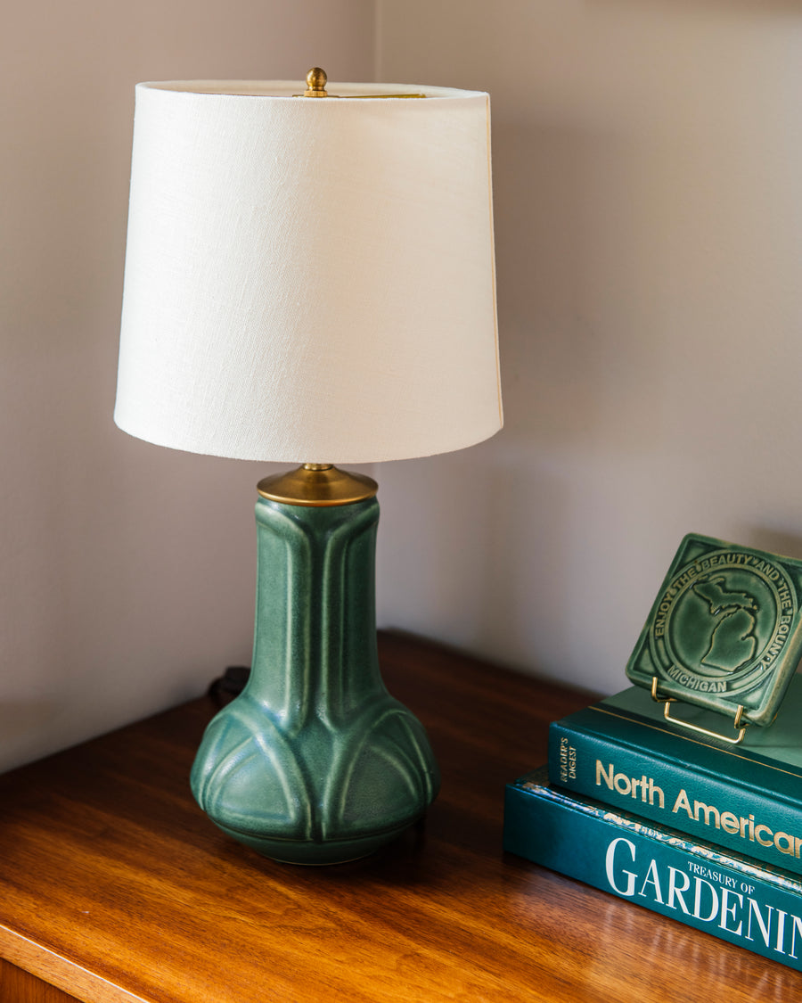 A matte green Elm glazed table lamp with an off-white shade next to a stack of books against a neutral wall.