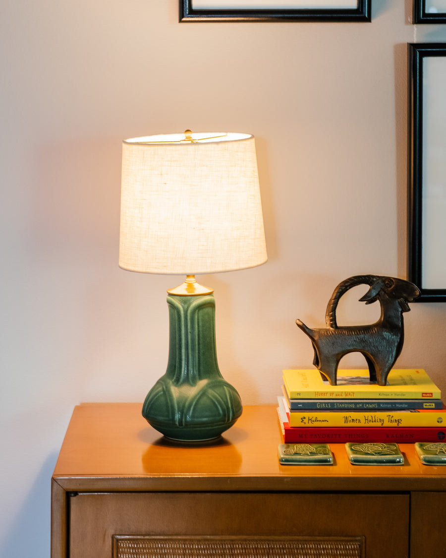 A matte green Elm glazed table lamp with an off-white shade next to a stack of books against a neutral wall with a metal goat statue. 