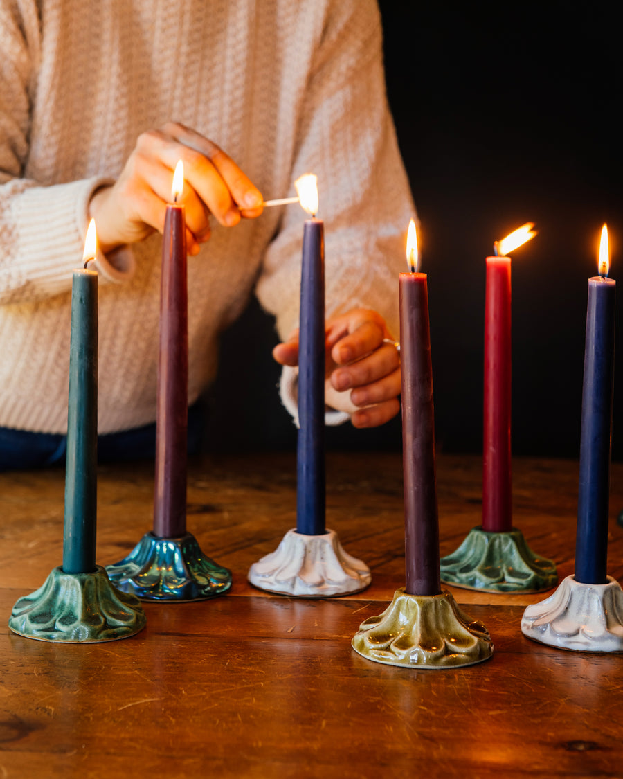 A person in a cream sweater is lighting a row of colorful candles on a wooden surface. The candles are held by decorative, scallopped, floral, ceramic candleholders crafted at Pewabic Pottery in a range of glaze color options from glossy "Honey", matte "Birch" and "Elm", to a semi-reflective "Matte Green Iridescent." 