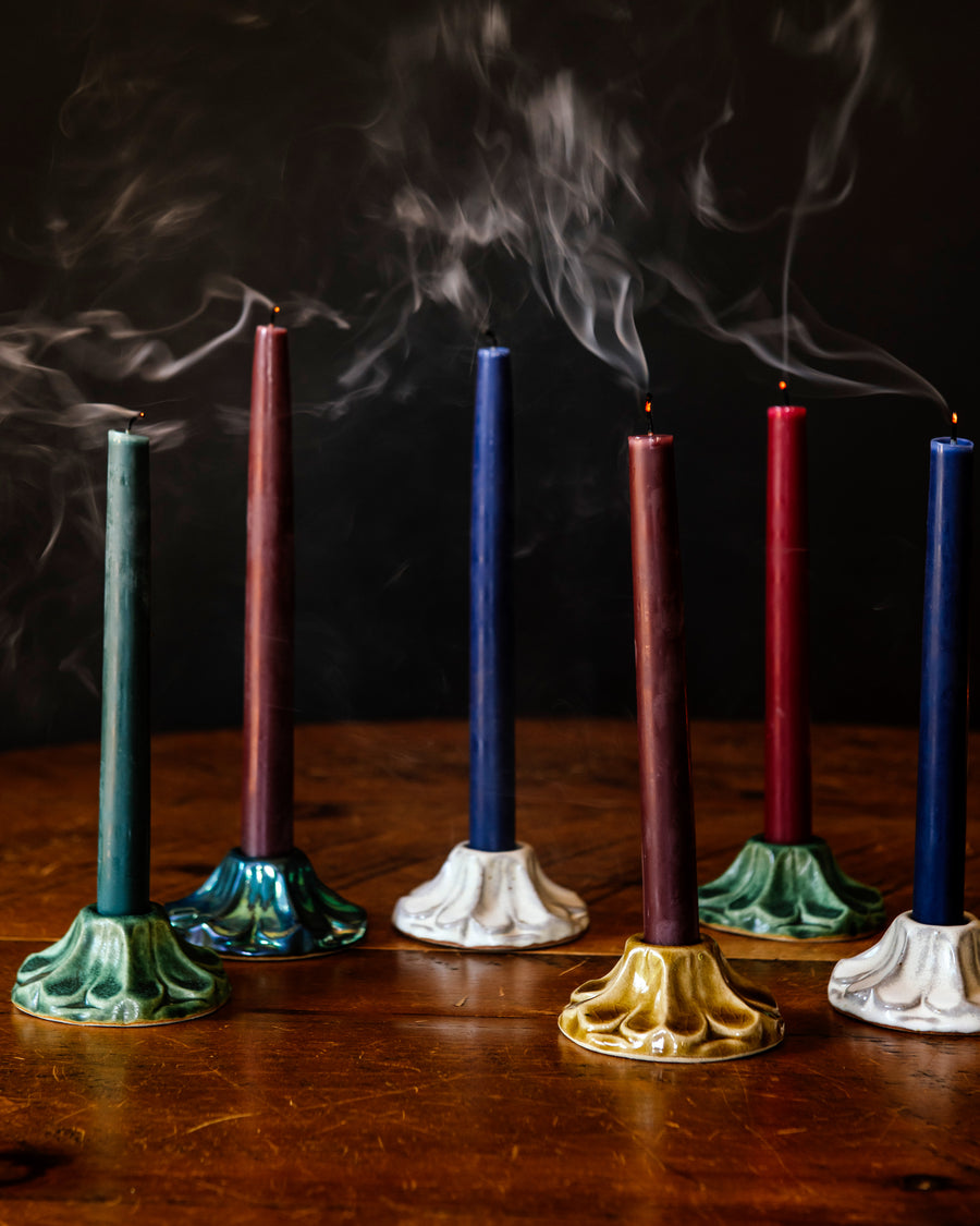 Set of colorful candles that have been recently blown out on a wooden surface against a dark background. The candles are held by decorative, scallopped, floral, ceramic candleholders crafted at Pewabic Pottery in a range of glaze color options from glossy "Honey", matte "Birch" and "Elm", to a semi-reflective "Matte Green Iridescent." 