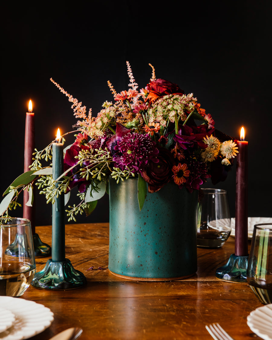 Floral arrangement in a freckled Greenstone vase on a wooden table with candles and glasses.