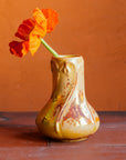 Delicate slip-casted snowdrop floral motifs dance across this Snowdrop Vase in a Sunstone Iridescent glaze that shimmers with the colors of a sunset. The texture of the vase varies with areas that are matte-yellow and reflective features glistening in orange and pinkish hues. The vase rests on terracotta roof tiles with a lime-wash orange background. A single orange poppy is proudly displayed inside of the vase. 