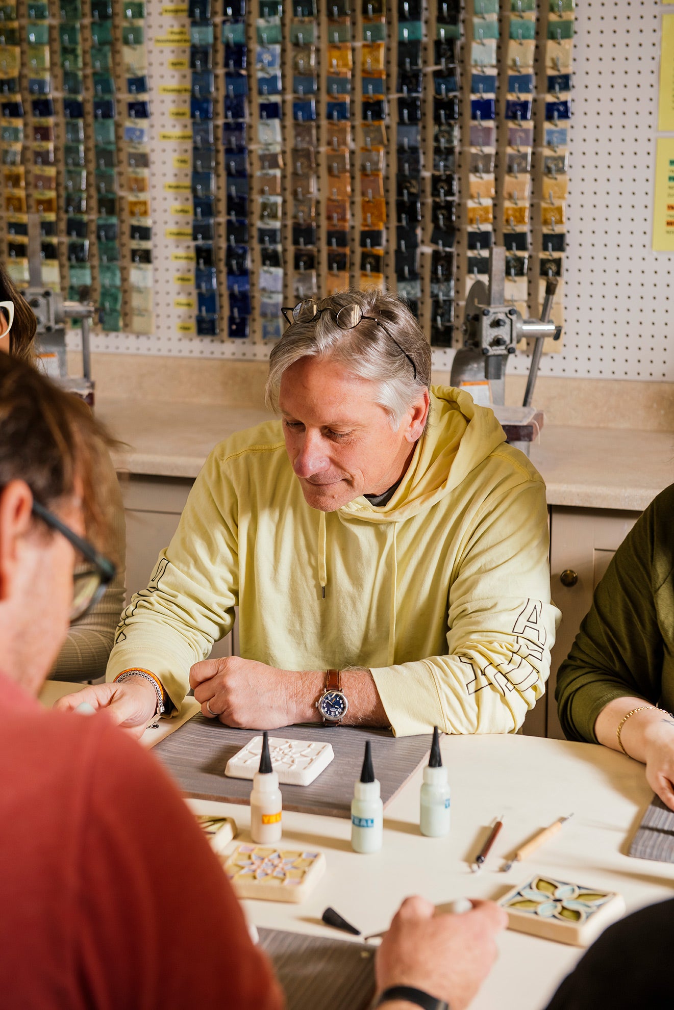 Man in a yellow sweatshirt sitting at a table with others, glazing a tile in the Pewabic Education Studio.
