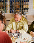 Man in a yellow sweatshirt sitting at a table with others, glazing a tile in the Pewabic Education Studio.