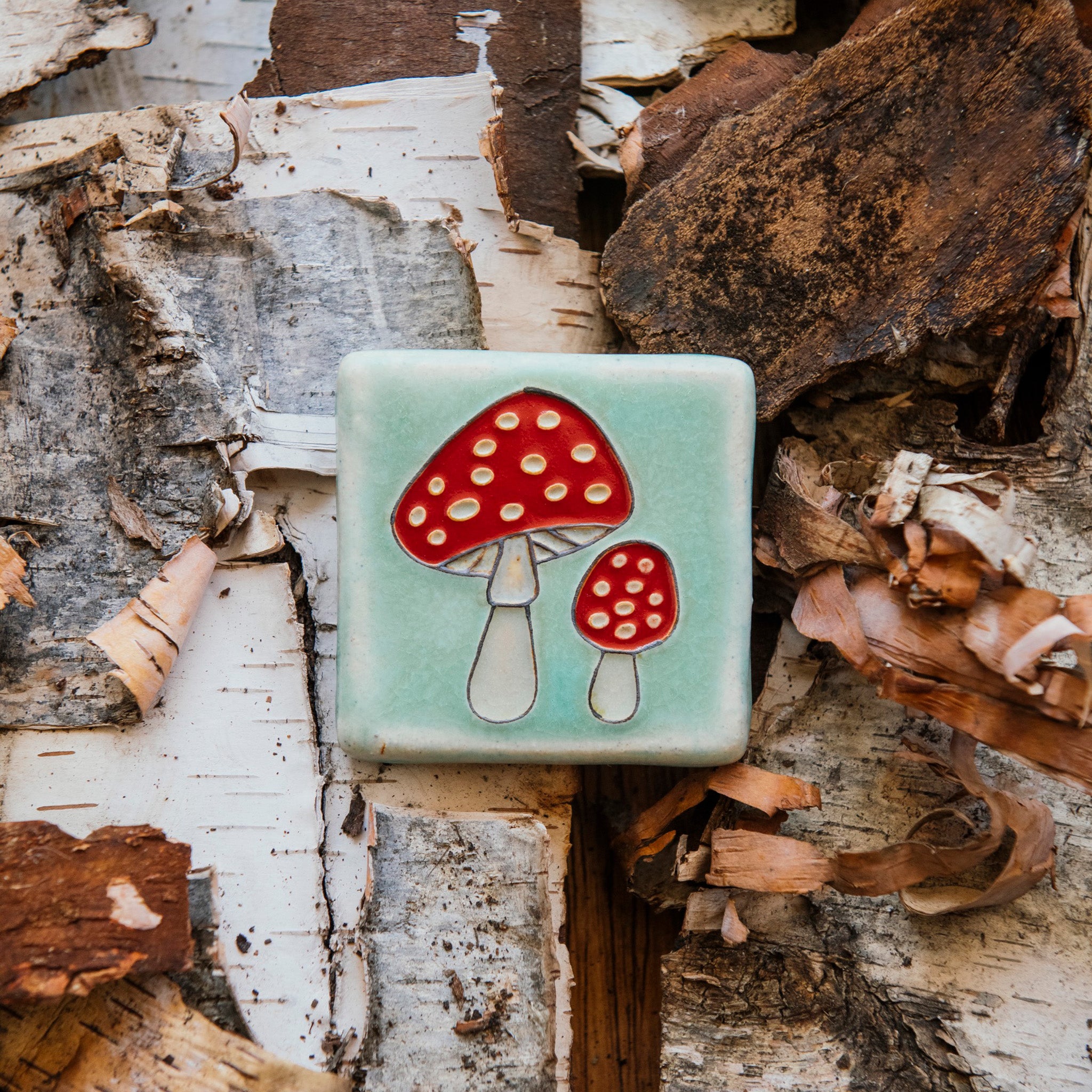 A hand-painted tile depicting a line drawing of a pair of Amanita mushrooms with classic red tops and white spots set on a light sea foam colored background glaze. 