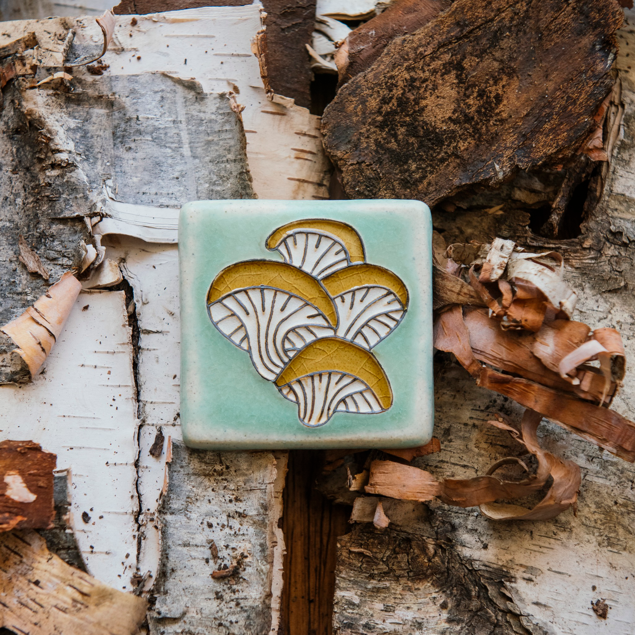 A hand-painted tile depicting a line drawing of Oyster mushrooms with golden yellow tops and white bases set on a light sea foam colored background glaze. 