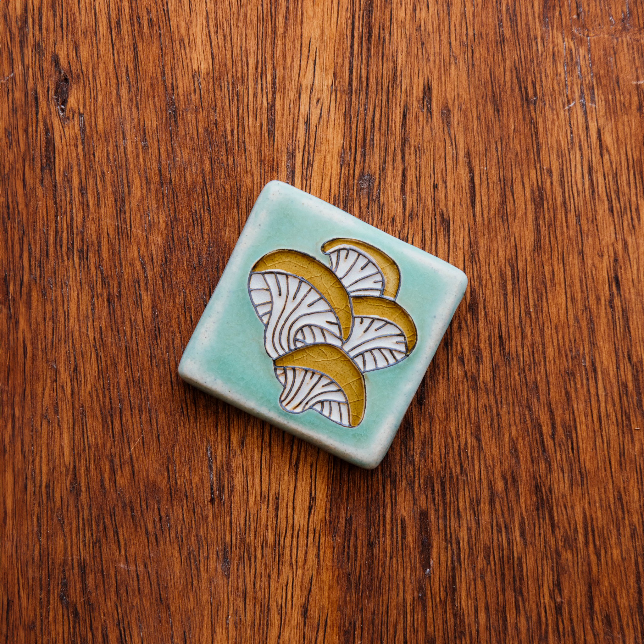 A hand-painted tile depicting a line drawing of Oyster mushrooms with golden yellow tops and white bases set on a light sea foam colored background glaze. 