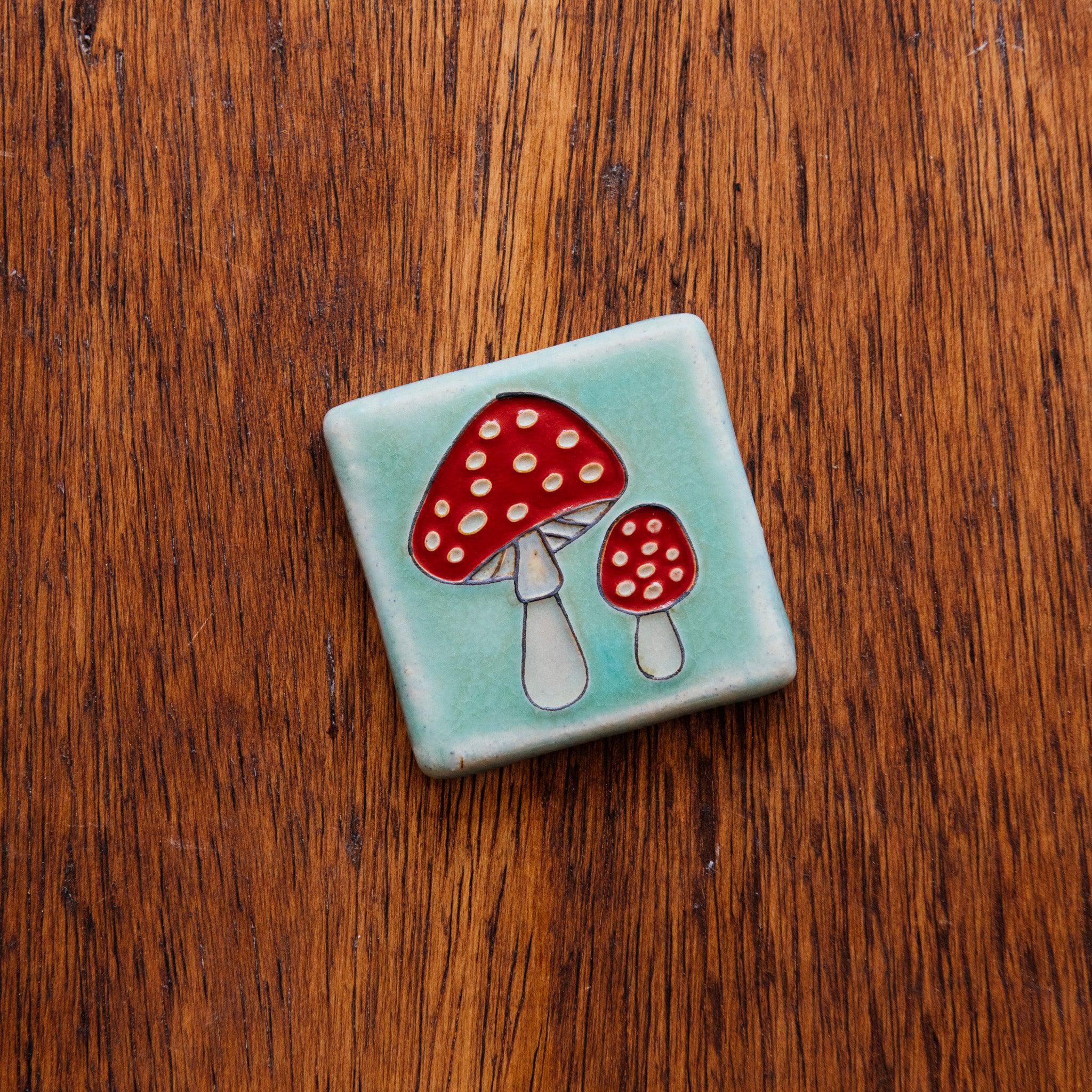 A hand-painted tile depicting a line drawing of a pair of Amanita mushrooms with classic red tops and white spots set on a light sea foam colored background glaze. 