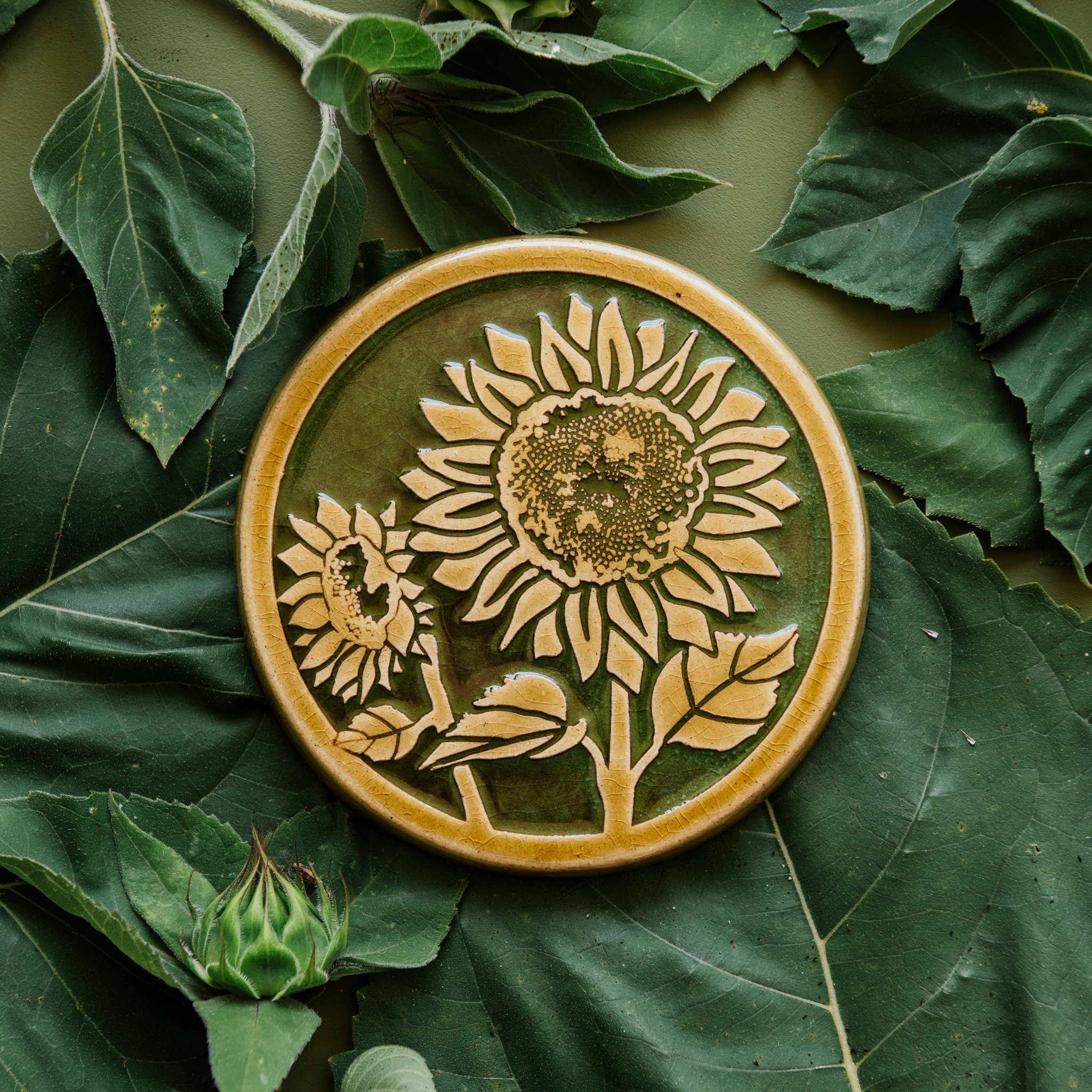 A two-tone glossy, round Sunflower Trivet with golden border and outline set against an emerald green background. 