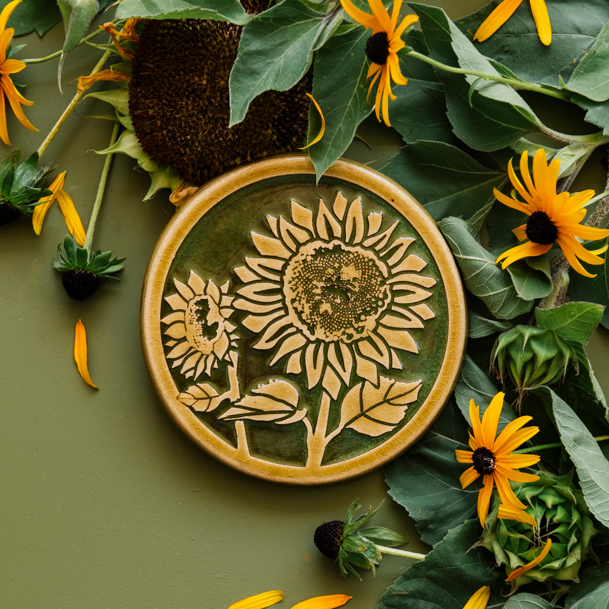 A two-tone glossy, round Sunflower Trivet with golden border and outline set against an emerald green background. 