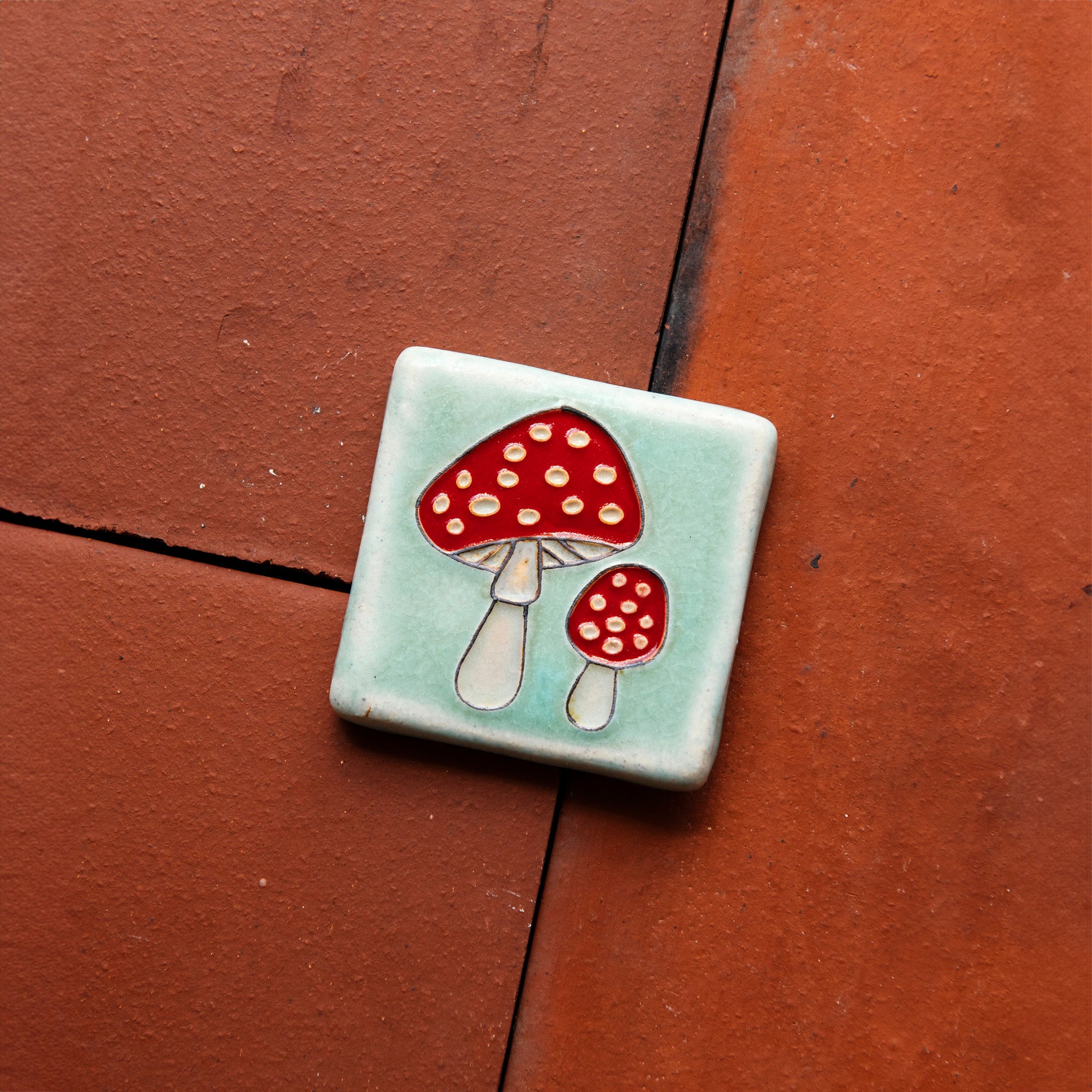 A hand-painted tile depicting a line drawing of a pair of Amanita mushrooms with classic red tops and white spots set on a light sea foam colored background glaze. 