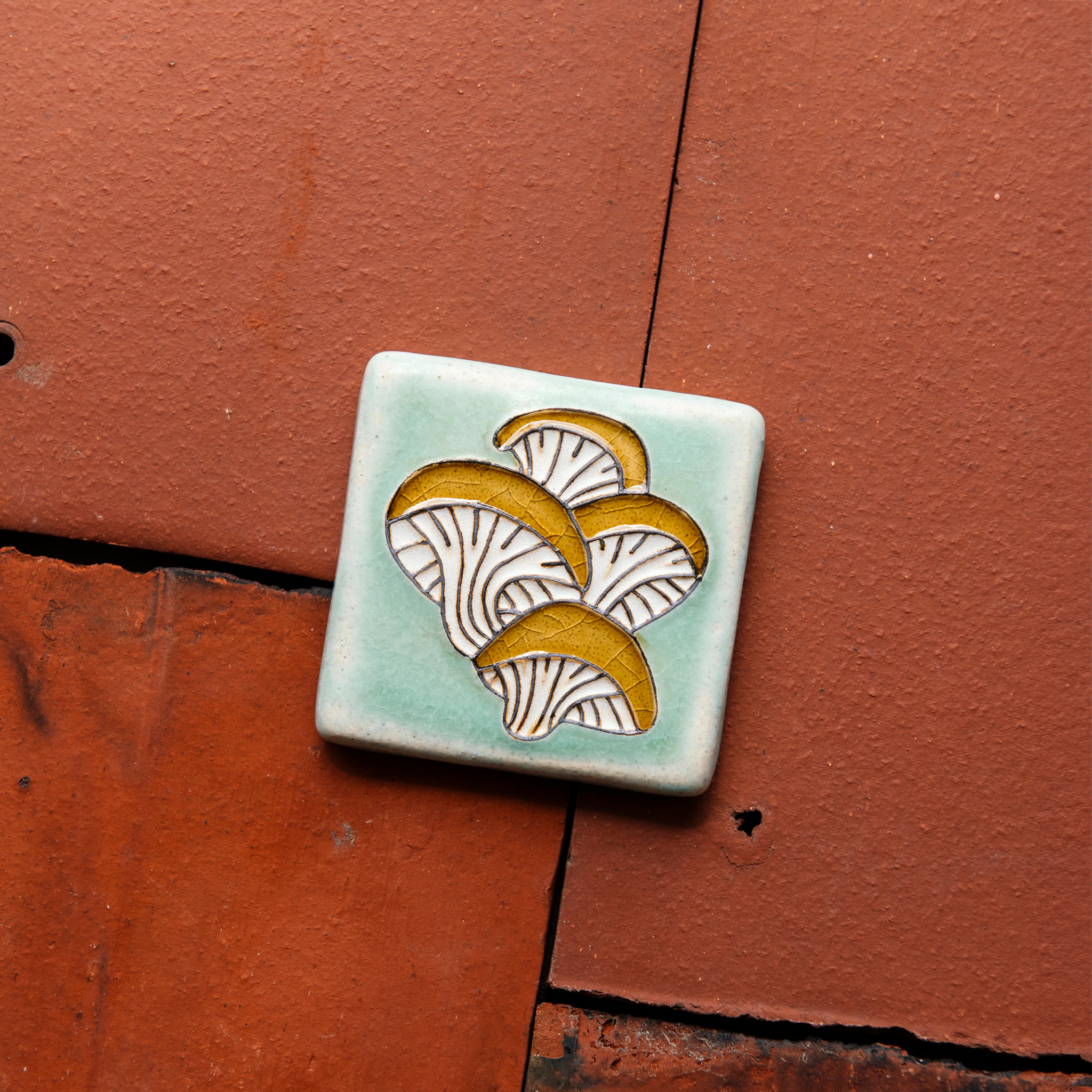A hand-painted tile depicting a line drawing of Oyster mushrooms with golden yellow tops and white bases set on a light sea foam colored background glaze. 