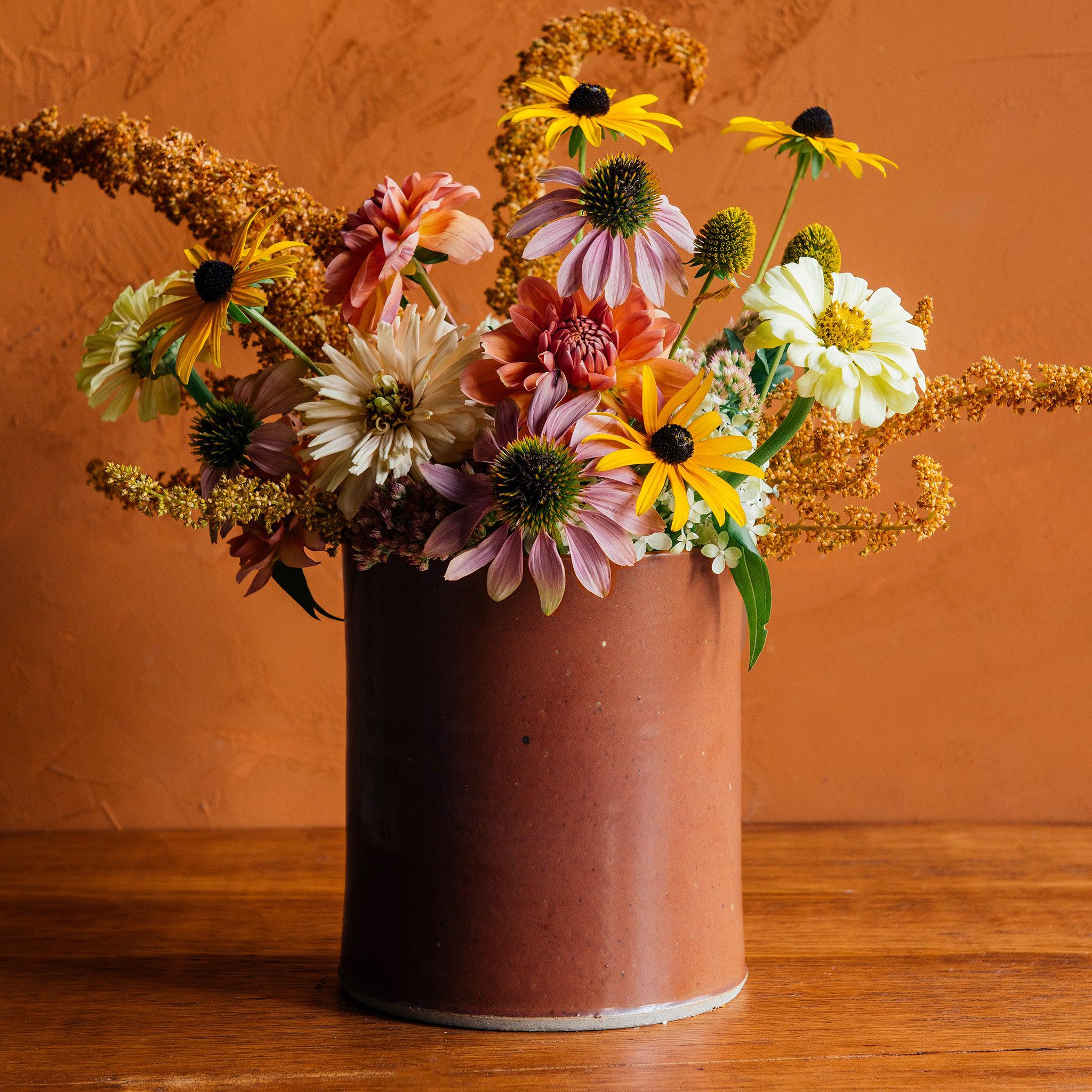 This Crock features the matte brownish-red tones of the Cinnamon glaze. Sitting on a wooden table, this crock is used as a vase, holding black eyed susans, dahlias and other end of summer blooms.