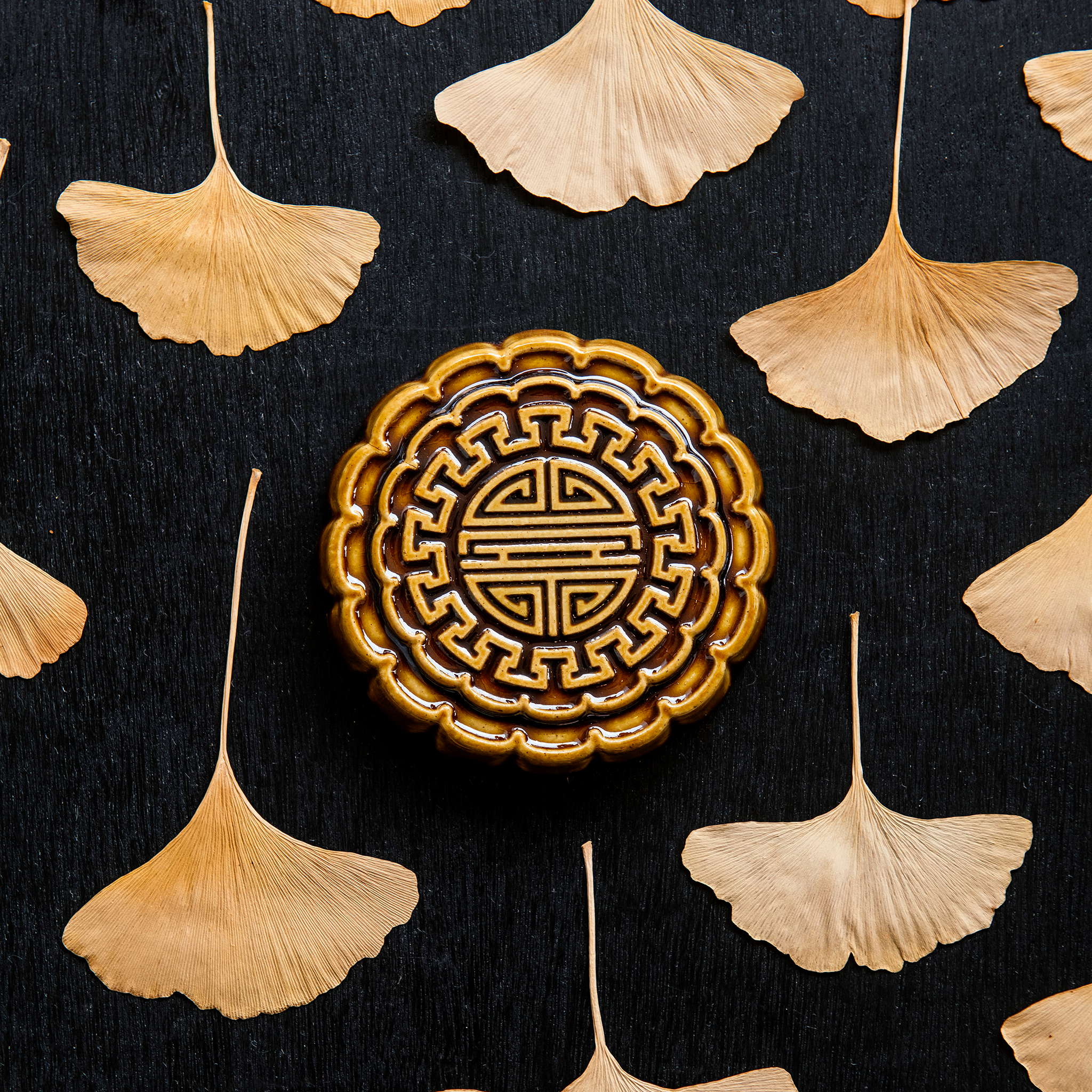 A Mooncake Paperweight in our glossy Molasses glaze, surrounded by dried ginkgo leaves.