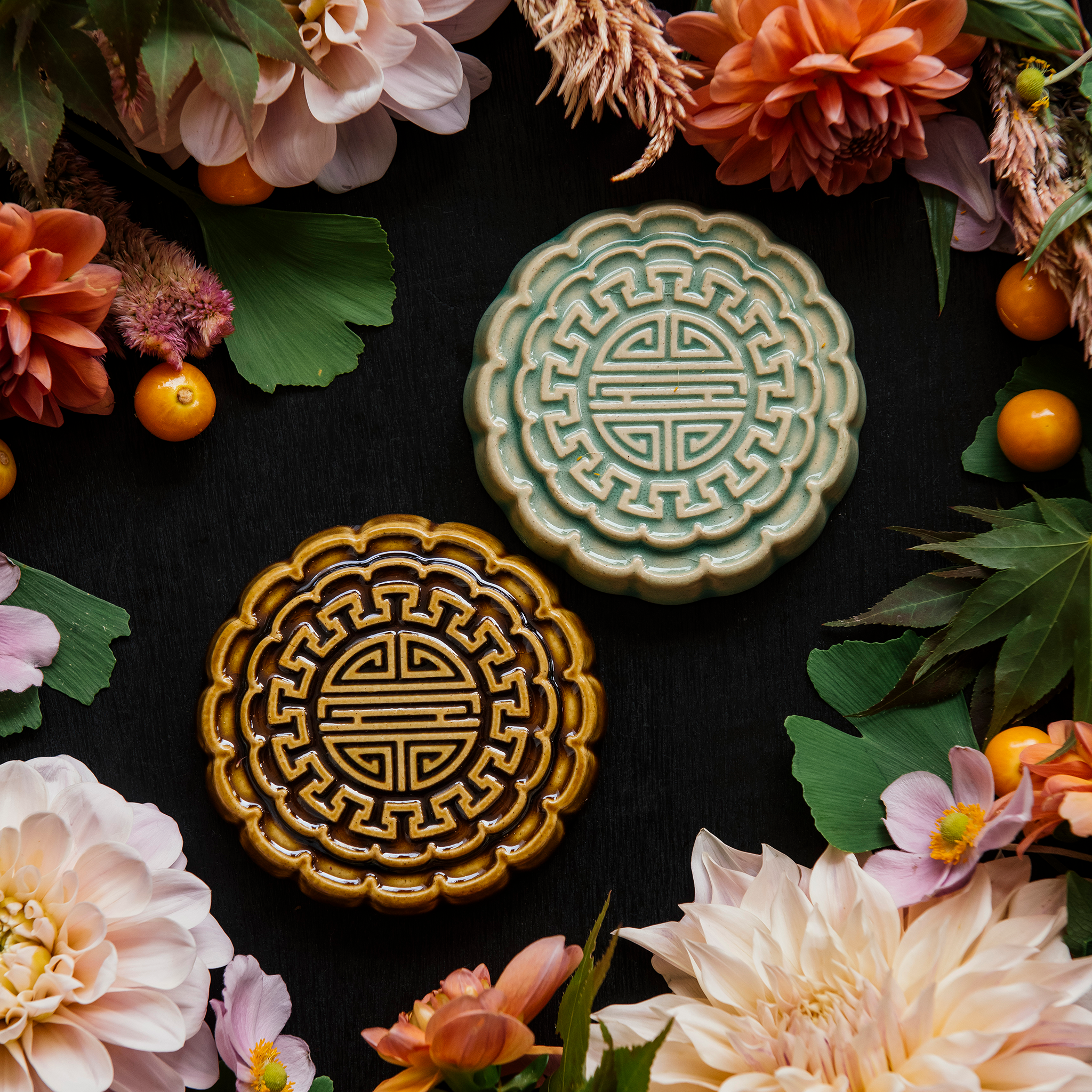 Two Mooncake Paperweights in glossy Celadon and Molasses glaze surrounded by bright, summer florals and gooseberries. 