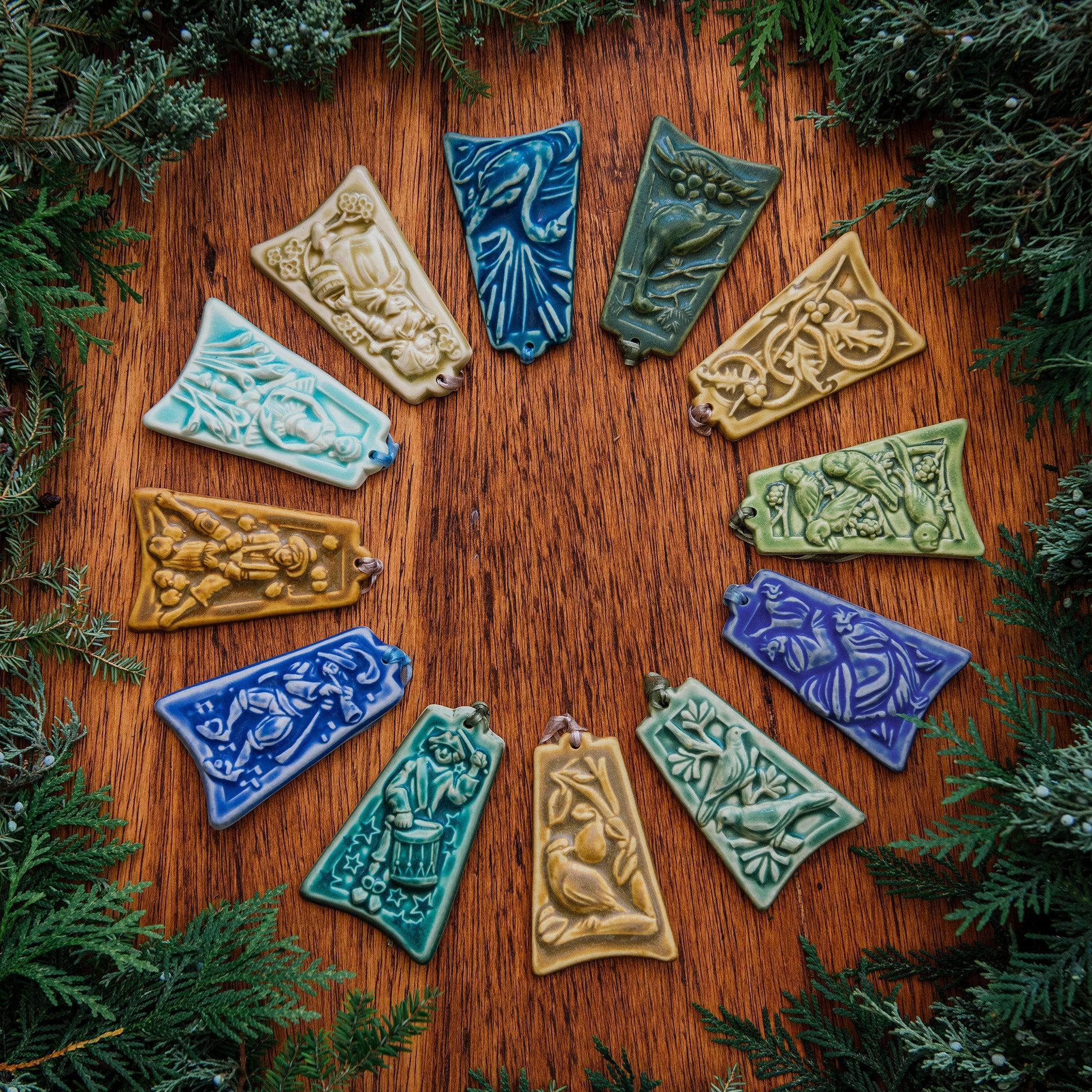 A colorful ceramic 12 Days of Christmas Ornament Set is splayed in a perfect circle with boughs of greenery surrounding them.