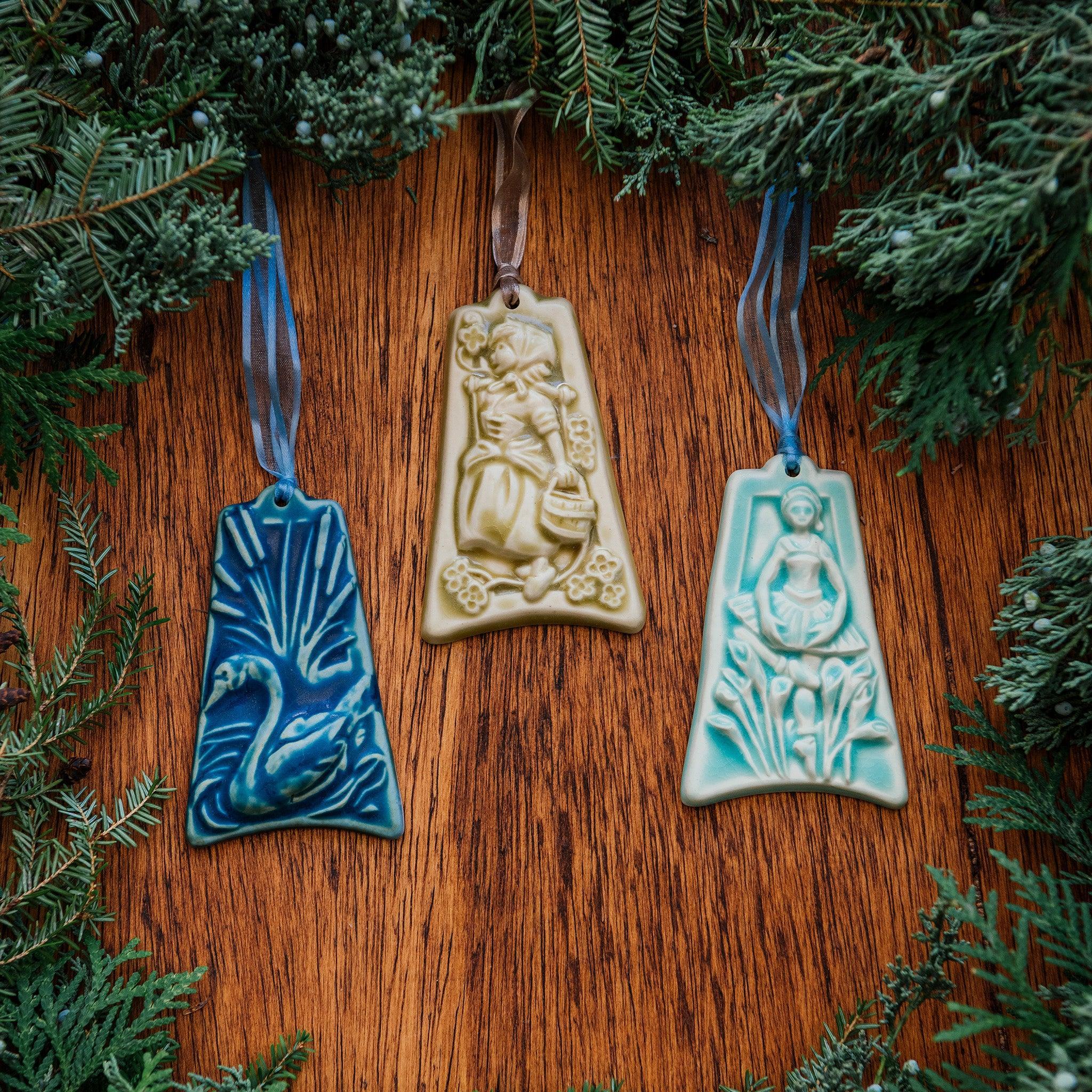 Three colorfully embossed ornaments are against a wood background with holiday greenery around them: one shows a swan swimming among seven cattails, another shows a young woman dressed as a milkmaid carrying a wooden bucket, the last has a ballerina mid-dance while nine calla lilies are being thrown to her.