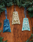 Three colorfully embossed ornaments are against a wood background with holiday greenery around them: one shows a swan swimming among seven cattails, another shows a young woman dressed as a milkmaid carrying a wooden bucket, the last has a ballerina mid-dance while nine calla lilies are being thrown to her.