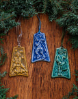 Three colorful embossed ceramic ornaments are on a wood backdrop with holiday greenery surrounding them: one shows a man jumping over a stump surrounded by fallen apples, one shows a man dressed in bloomers and a feathered hat as he plays a horn, the last one has a man dressed in Revolutionary War garb as he marches while playing a drum.  