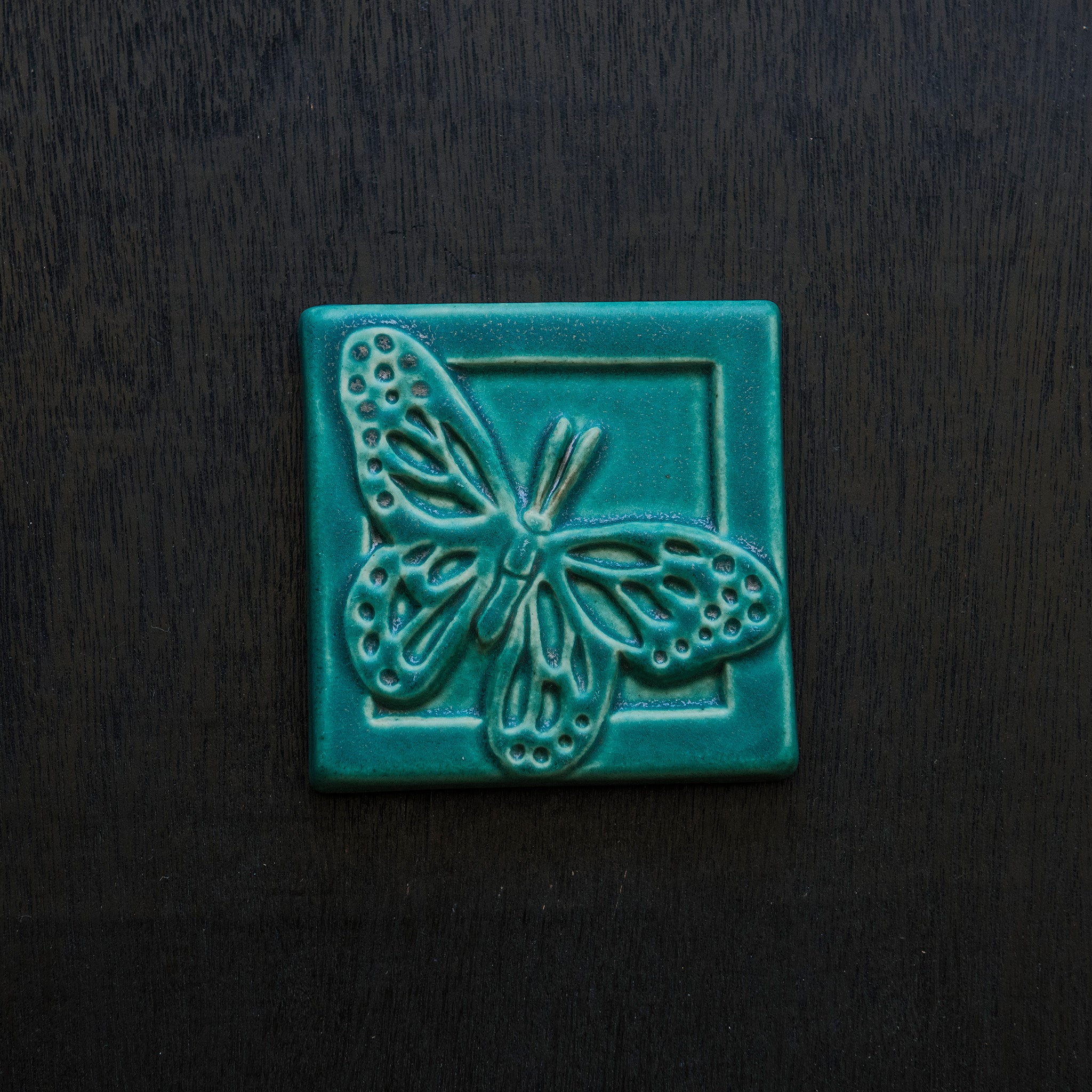 The ceramic Butterfly tile's design sits diagonally on the 4x4 square tile. Its wings are outstretched and it is floating above a depressed border that surrounds the tile. This gives the illusion that the butterfly is flying out toward you. This tile features the matte turquoise Pewabic Blue glaze.