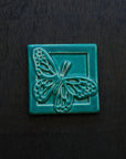 The ceramic Butterfly tile's design sits diagonally on the 4x4 square tile. Its wings are outstretched and it is floating above a depressed border that surrounds the tile. This gives the illusion that the butterfly is flying out toward you. This tile features the matte turquoise Pewabic Blue glaze.