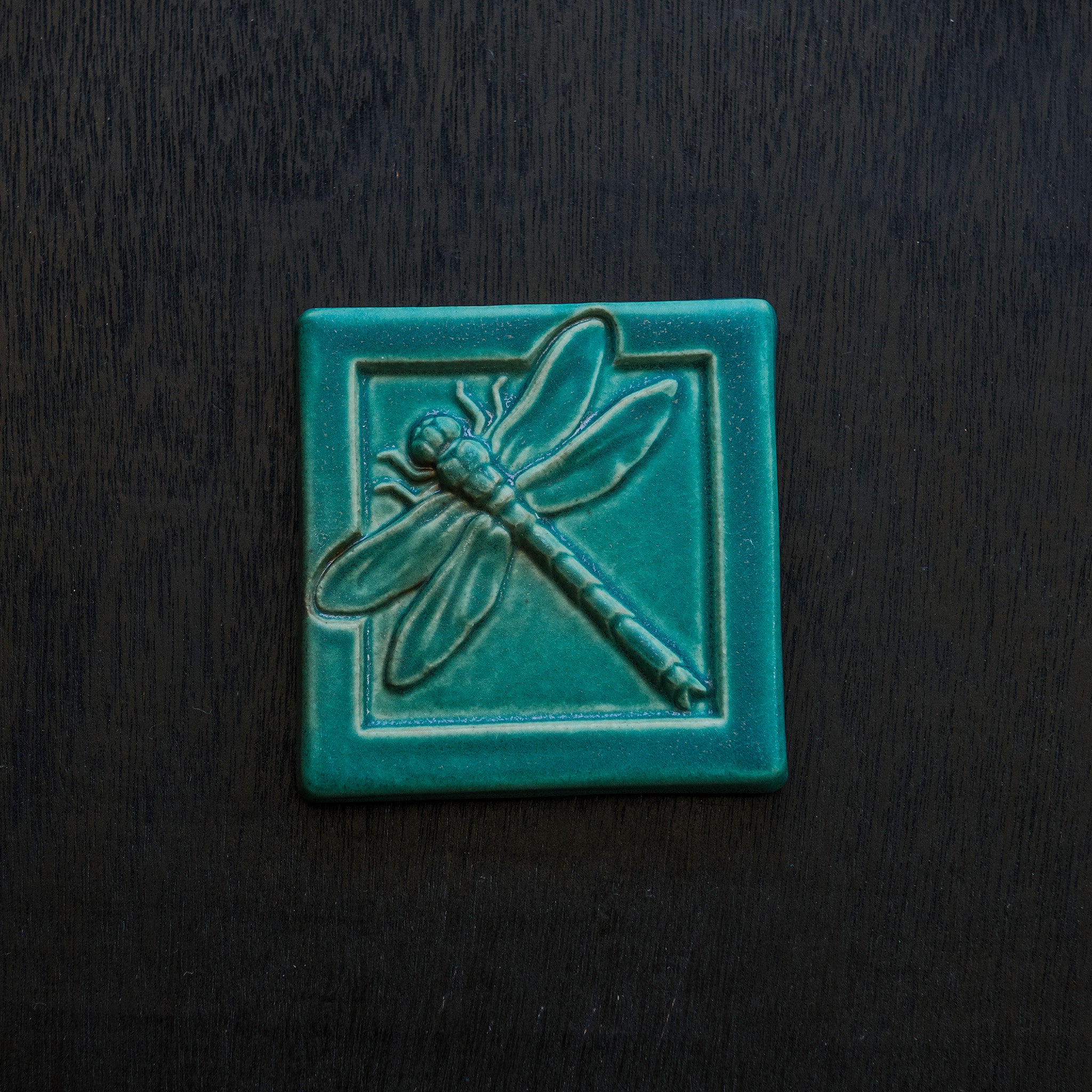 The Dragonfly Tile features a detailed dragonfly with wings outstretched. It is sitting horizontally on the square tile with a thick border around the edge. This Dragonfly Tile features the blueish-teal Pewabic Blue glaze.