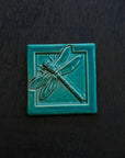 The Dragonfly Tile features a detailed dragonfly with wings outstretched. It is sitting horizontally on the square tile with a thick border around the edge. This Dragonfly Tile features the blueish-teal Pewabic Blue glaze.