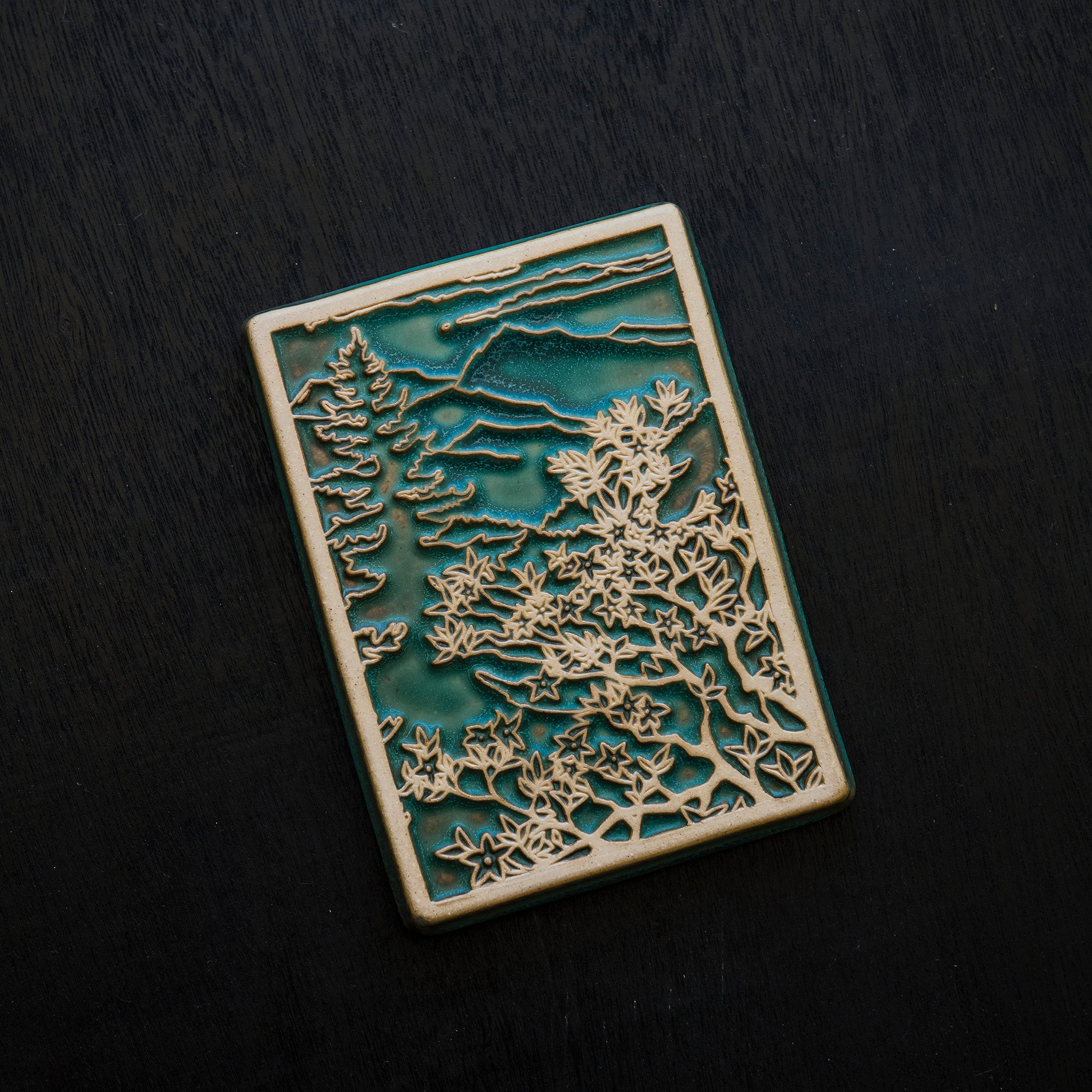 This Mountain View Tile features a a large flowery bush in the foreground with a tall pine tree behind it. In the distance, craggy mountains sit under a cloudy sky.  