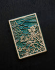 This Mountain View Tile features a a large flowery bush in the foreground with a tall pine tree behind it. In the distance, craggy mountains sit under a cloudy sky.  