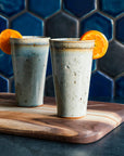 Two ceramic Pewabic Pints in a glossy Frost glaze, garnished with orange slices on a wooden cutting board against a blue hex tile background. 