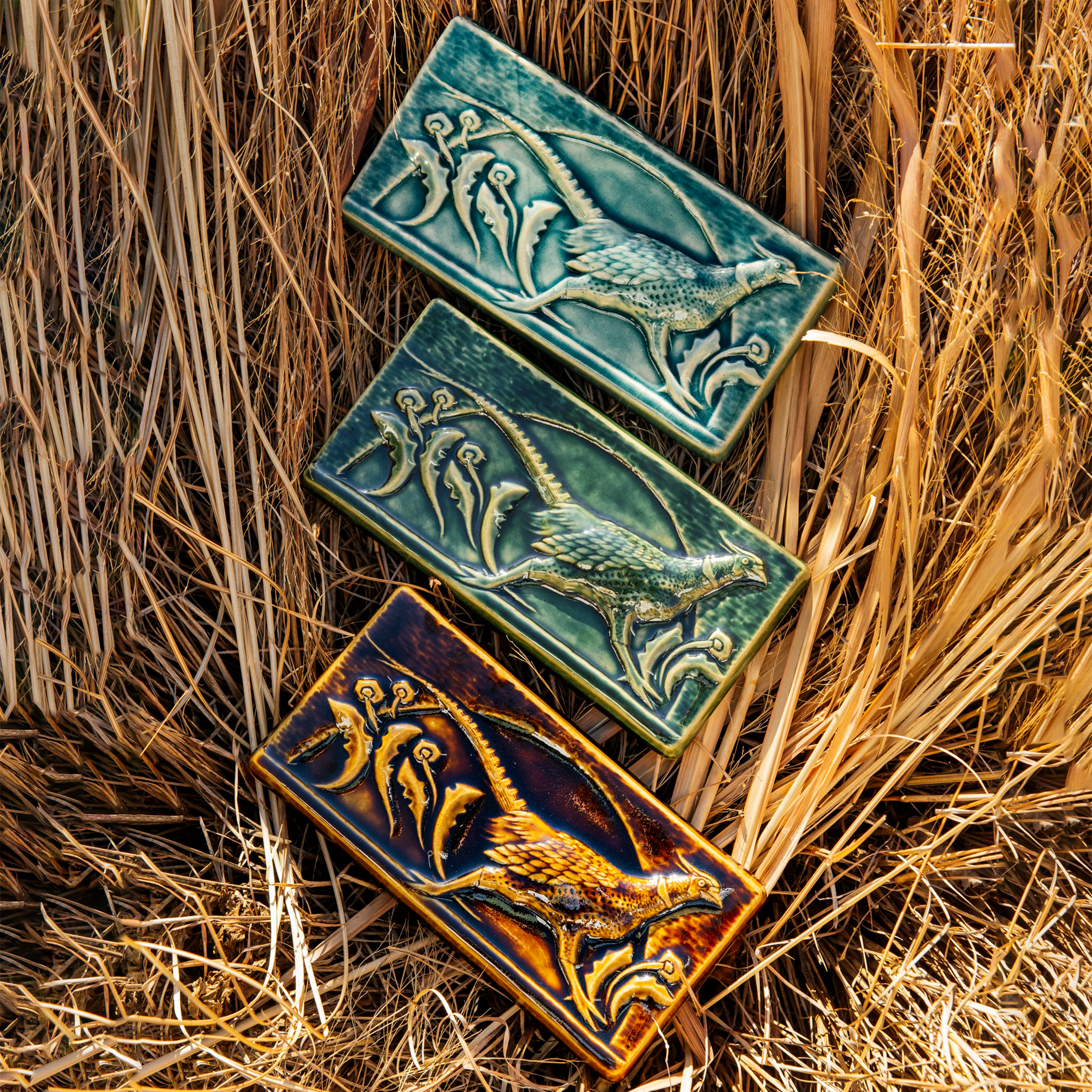 Three Pheasant tiles rest on a bed of brown grasses. The tiles are in each glaze option - Kale, Glacier Gloss and Molasses.