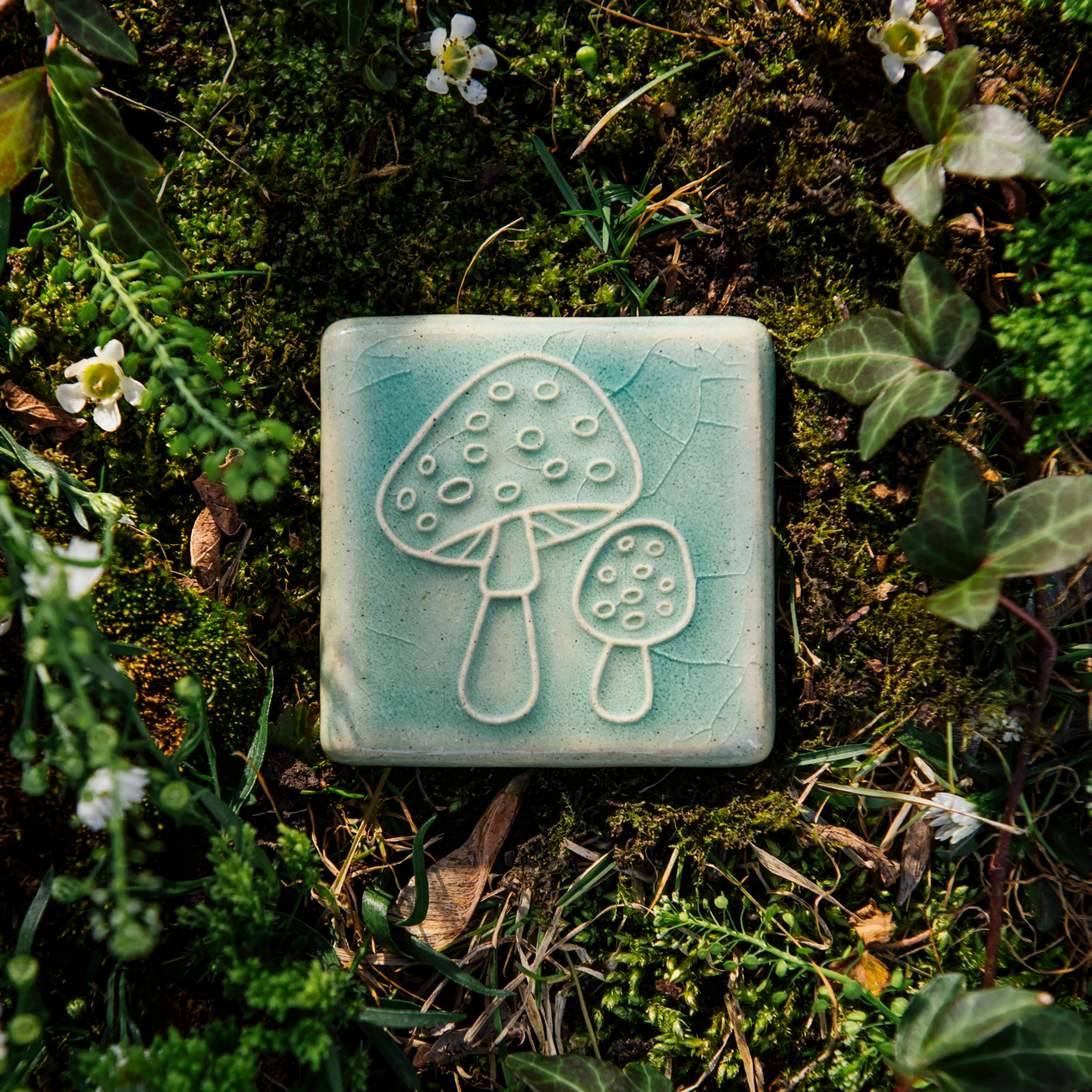 The Amanita Mushroom Tile features a line-drawing of two mushrooms, one small and one large. Each mushroom has polka dot-like circles on its cap and the the larger mushroom is slightly angled, showing off the gills that peek out under its round top.
