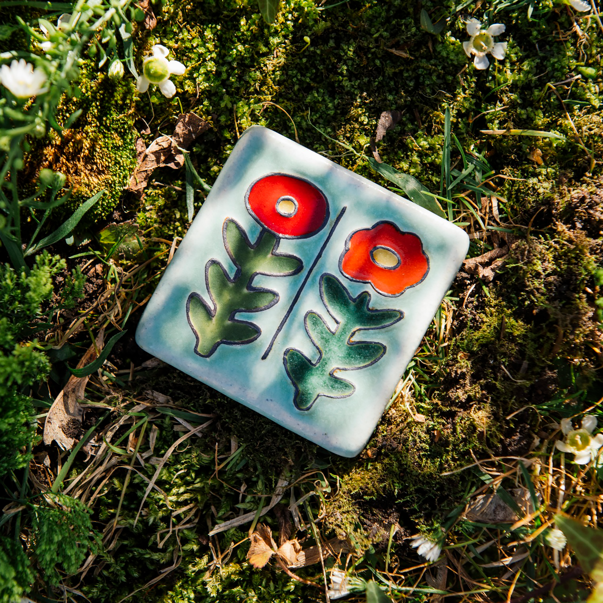 This handpainted tile in the Poppy Palette features bight red and deep orange petals and medium green stems. The background of the tile is a pale blue color.