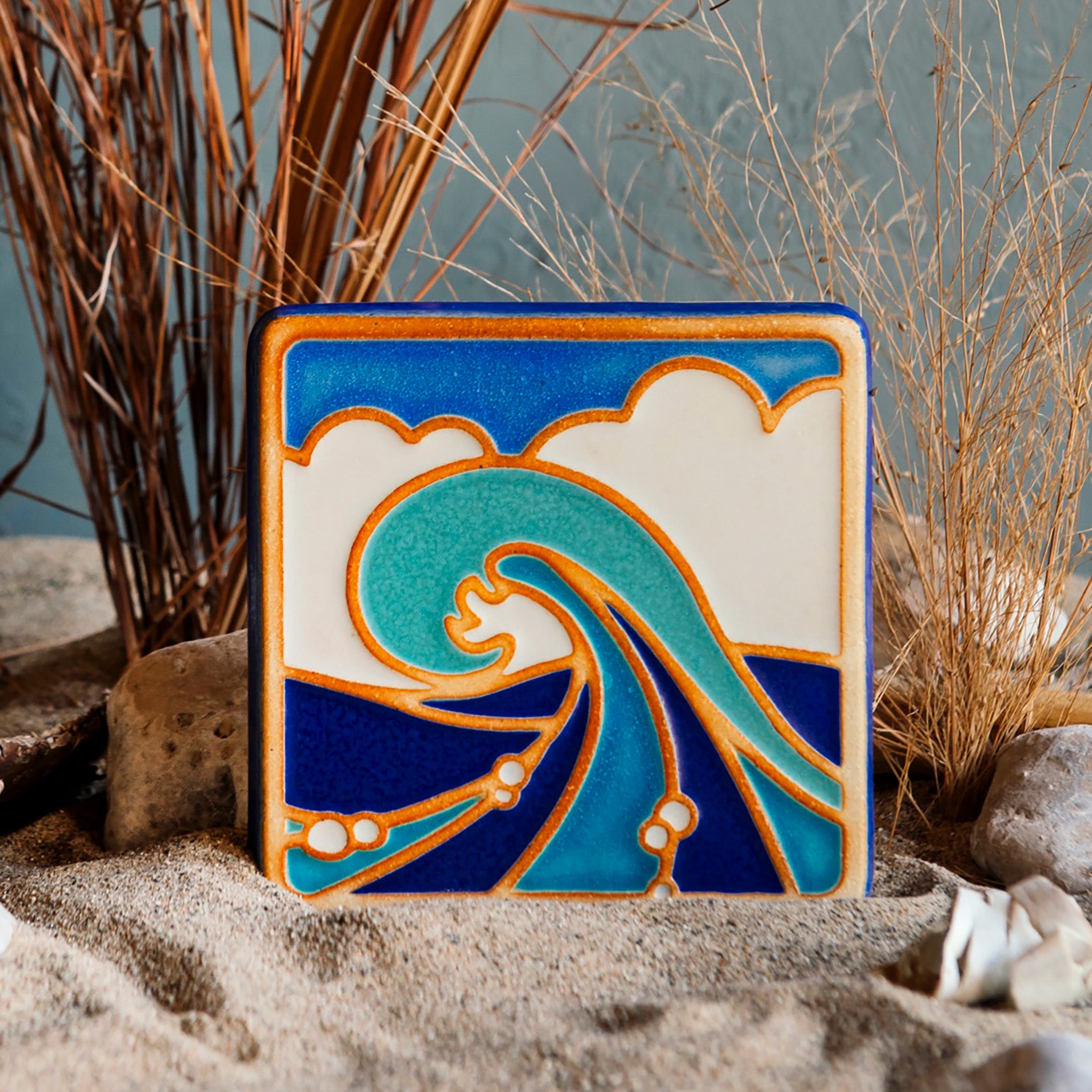 The Wave Tile features a line drawing of a large, curled wave. There are bubbles scattered around its base with a huge puffy cloud behind it that almost covers the bright blue sky. The hand painted tile has multiple blue hues with bright white accents.