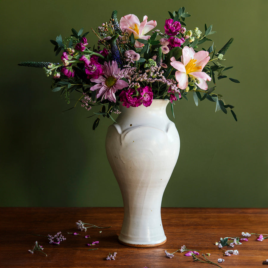 Our Large Classic Vase in a creamy white Birch glaze with brown, iron speckles throughout. The vase is set against a deep, olive-green backdrop with a warm wood foreground. Bright summer flowers decorate the vessel and include a range of purples, soft-pinks, and muted magentas.