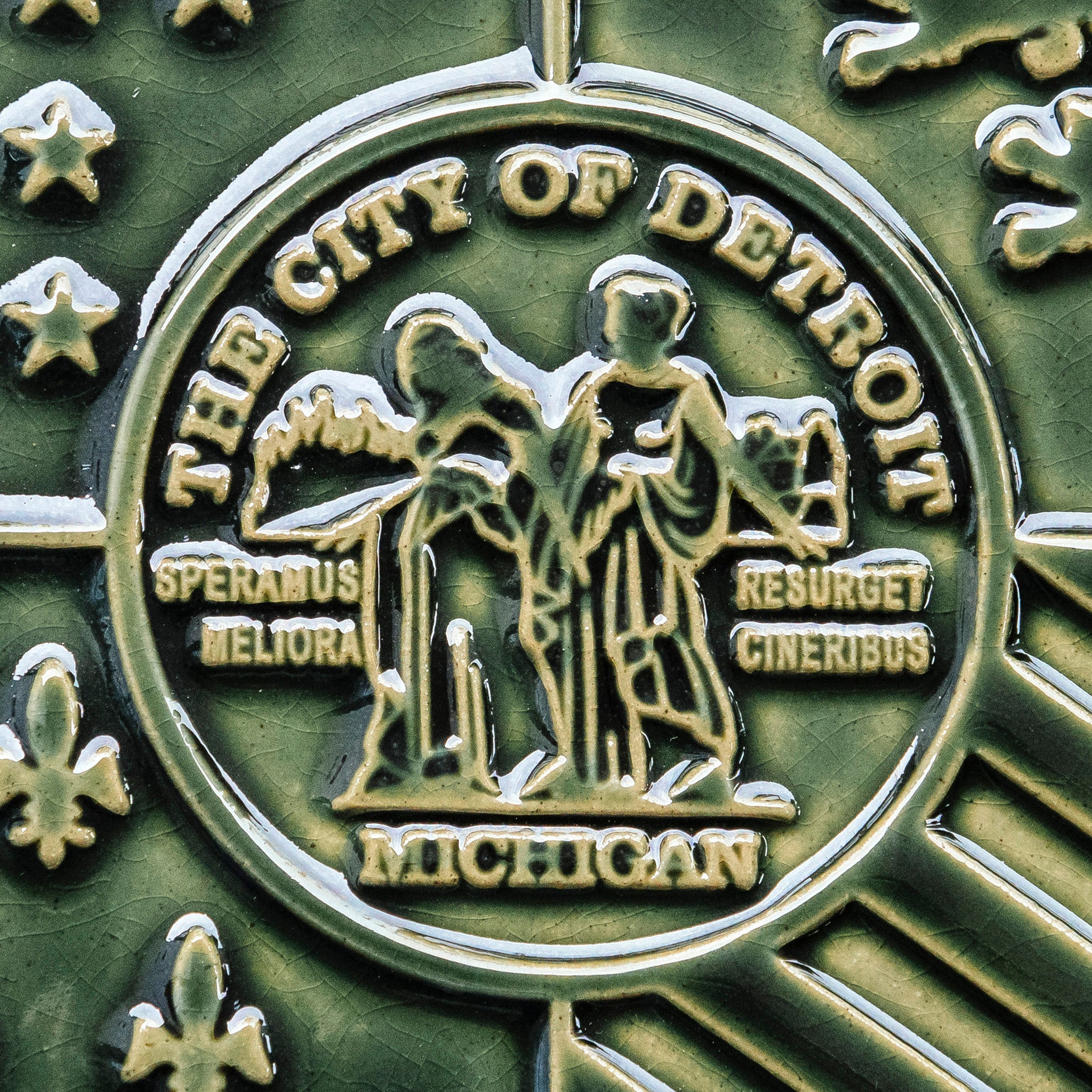 Detail photo of the centerpiece of the Detroit Flag Tile. The circular emblem reads “The City of Detroit” and “Michigan” at the top and bottom. At its center, are two women in red and blue robes. Fire is in the background of the woman on the left and reads “Speramus Meliora” which is latin for “We hope for better things”. The woman on the left is surrounded by a cityscape and reads “Resurget Cineribus” which is “It will rise from the ashes” in latin. 