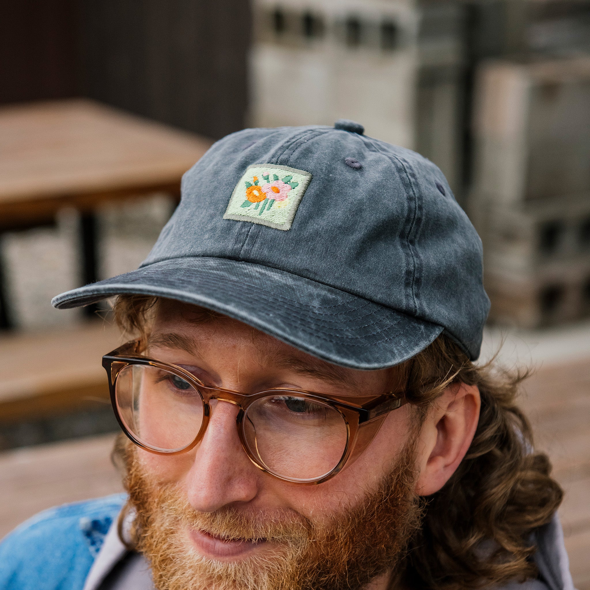 Man wearing a dark gray cap with a square colorful floral patch in the pottery courtyard.