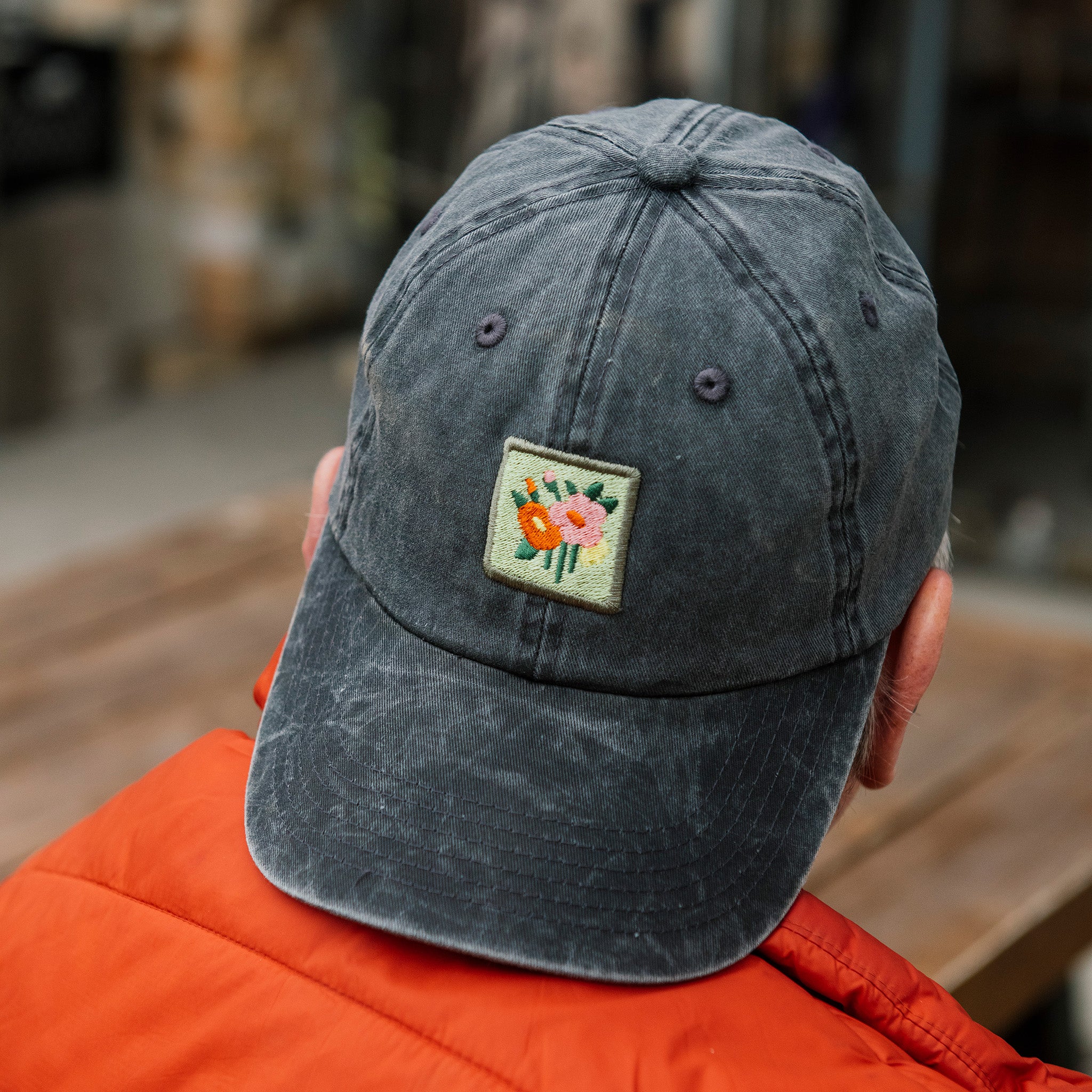 Person wearing a dark gray cap with a floral patch, sitting outdoors.