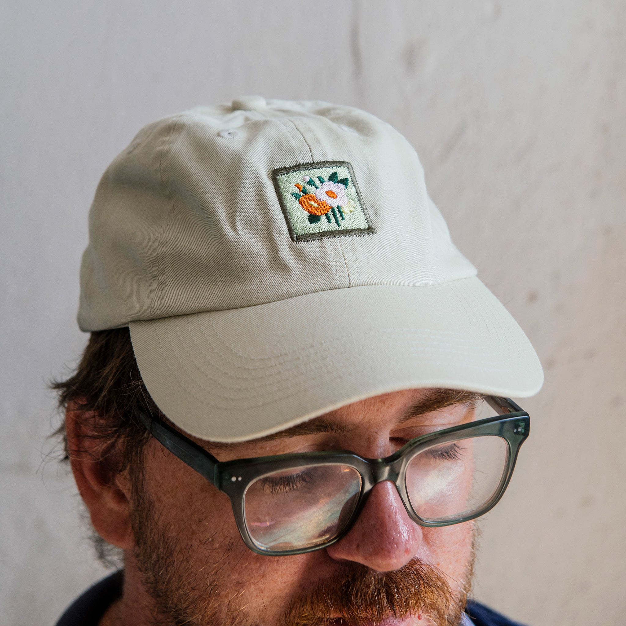 A man wears the off-white version of the Floral Tile Hat. The colorful, square patch is embroidered onto the muted white baseball cap.